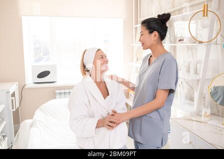 Zufriedene reife Beauty Salon Gast in Robe sitzen auf Verfahren Tisch und Hand der asiatischen Kosmetologen halten, während ihr nach dem Verfahren danken Stockfoto