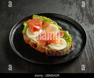Scheibe Brot mit gekochtem Ei und Lachs auf schwarzem Teller Stockfoto