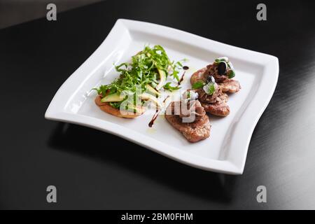 Blick von oben auf den weißen Teller mit leckeren Bruschettas mit Salatrakete und Avocado im Restaurant. Drei kleine, mit Paste verzierte Rindersteaks, Stockfoto
