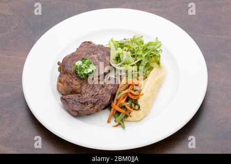 Ein geröstetes Steak mit Kartoffelpüree, gebratenem Gemüse und Salat in einem weißen Keramikplatte auf einem Holztisch Stockfoto