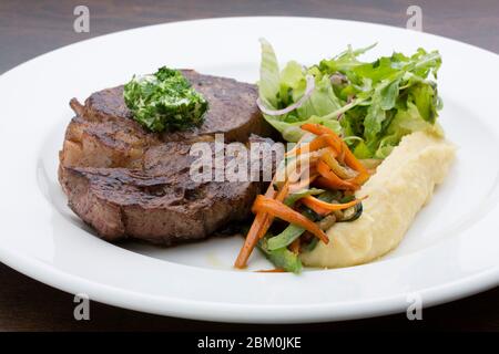 Ein geröstetes Steak mit Kartoffelpüree, gebratenem Gemüse und Salat in einem weißen Keramikplatte auf einem Holztisch Stockfoto