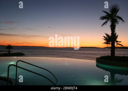 Ein Schwimmbad und eine Palme mit dem Meer und dem Sonnenuntergang im Hintergrund Stockfoto