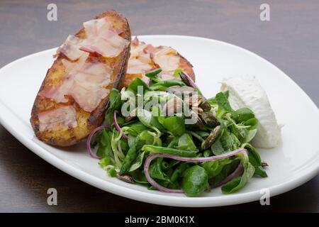 Gebackene Kartoffeln mit Speck, frischem Käse und einem Salat in einem Keramikplatte auf einem Holztisch Stockfoto