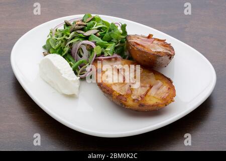 Gebackene Kartoffeln mit Speck, frischem Käse und einem Salat in einem Keramikplatte auf einem Holztisch Stockfoto