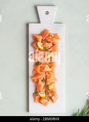 Scheiben von hausgemachtem geräuchertem Lachsfilet mit Dill und Zitrone auf Marmorschneidebrett auf hellem Hintergrund serviert. Draufsicht. Gesunde Ernährung Stockfoto