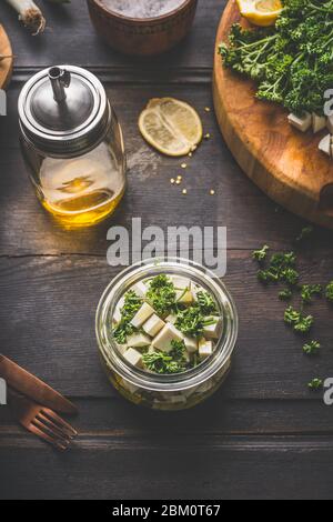 Feta-Käsewürfel Glas mit Olivenöl und Kräutern auf dunklem rustikalem Hintergrund. Marinierter Feta-Käse. Hausgemachte Speisen. Landhausstil. Draufsicht Stockfoto