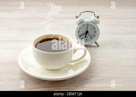 Morgenkaffee schwarz heißen Espresso mit Dampf in weißer Tasse, Untertasse auf Holztisch mit Uhr. Stockfoto