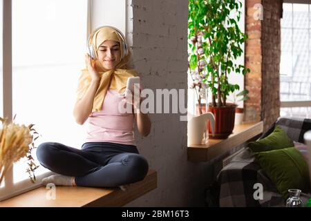 Eine hübsche junge muslimin zu Hause während der Quarantäne und Selbstisolierung. Arabisches weibliches Modell, das Zeit nützlich ist, isoliert zu sein. Konzept der Gesundheitsversorgung, Kommunikation, Bildung während der Pandemie. Stockfoto