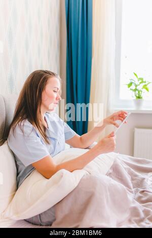 Junge schöne Frau sitzt auf einem Bett unter einer Decke und lesen Buch auf einer Lasche in einem blauen Pyjama. Stockfoto