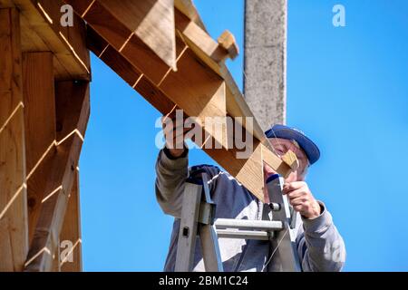 Ältere grauhaarige Baumeister sammelt den Rahmen eines hölzernen Landhaus stehend auf der Treppe gegen den blauen Himmel. Die körperliche Aktivität der elde Stockfoto