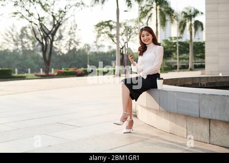 Schöne junge asiatische Frau, die ihr Smartphone anschaut und im urbanen Hintergrund lächelt Stockfoto