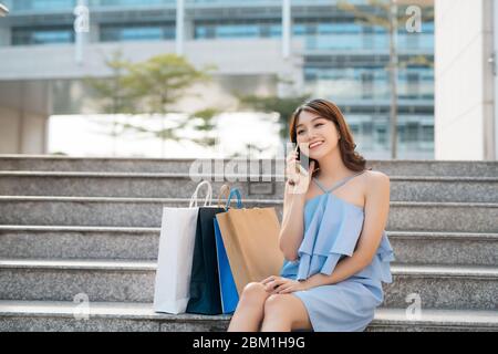 Fröhliche junge Asiatin mit Einkaufstaschen, die auf dem Handy telefonieren und auf einer Treppe sitzen. Stockfoto