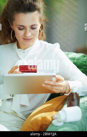 Elegante Mittelalterhausfrau in weißer Bluse mit Kreditkarte und leere Medikamentenflasche Kauf Apotheke online mit Tablet-PC in modernen Hause in sonnig Stockfoto