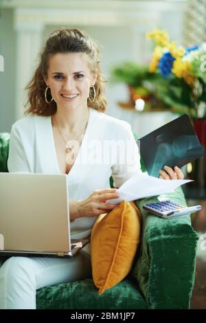 Portrait of happy trendy weiblich in weiß Bluse mit Dokumenten und Taschenrechner Wahl der Krankenversicherung digital in modernen Hause an sonnigen Tag. Stockfoto