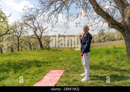 Sportlicher alter Mann macht Übungen mit Hanteln. Foto von gesunde schöne Active man mit Fit Muskelkörper unter blühenden Garten. Stockfoto