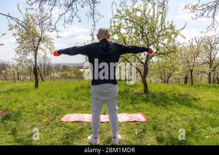 Älterer Mann in Sportkleidung, der sich wieder an die Kamera hält, hält rote Hanteln in den Händen, während er im blühenden Garten trainiert Stockfoto