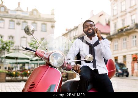 Schöner bärtiger afrikanischer Geschäftsmann genießt seine erfolgreiche Telefongespräche, während er auf dem modernen roten Motorrad auf dem Hintergrund von alt sitzt Stockfoto