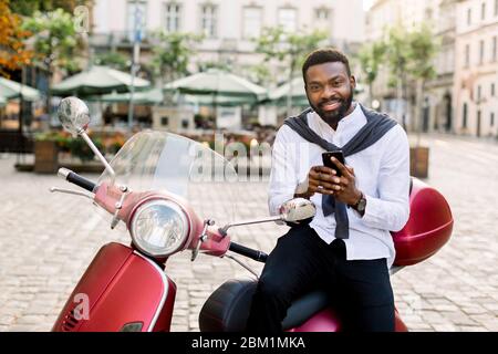Porträt des modischen afrikanischen Geschäftsmann trägt weißes Hemd und schwarze Hosen, sitzt auf dem roten Roller in der Stadt, lächelnd und Blick auf die Kamera Stockfoto