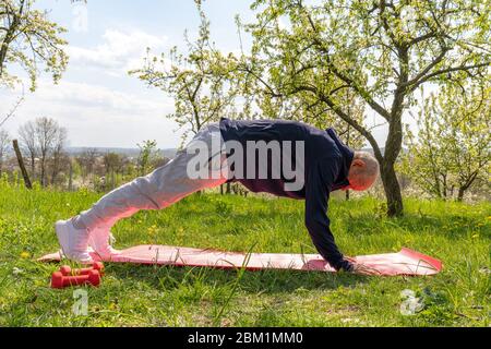 Motivierter und konzentrierter Senior Pencioneer mit schwarzem Running Outfit, das in Plankenposition auf einer Yogamatte inmitten eines blühenden Gartens steht Stockfoto