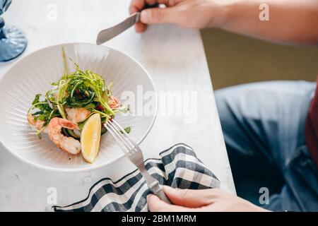 Salat mit Garnelen, Rucola, Zitrone, Tomaten in tiefer Schüssel, mit Händen von Männern, hält Besteck Gabel. Heller Hintergrund. Draufsicht. Stockfoto