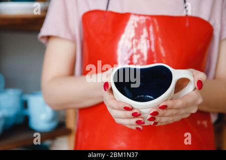 Junge Frau in roter Schürze für Keramik hält Herz-förmigen Becher. Stockfoto