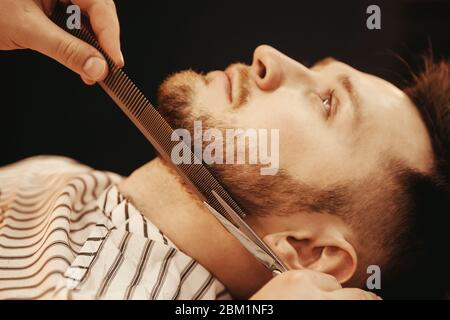 Trimmen des Mannbarts. Hände des Barbers halten Schere und Kamm. Dunkler Hintergrund Stockfoto