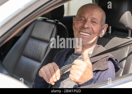 Ein alter glücklicher Mann mit kabellosen Kopfhörern, der ein Auto fährt und die Daumen nach oben zeigt Stockfoto