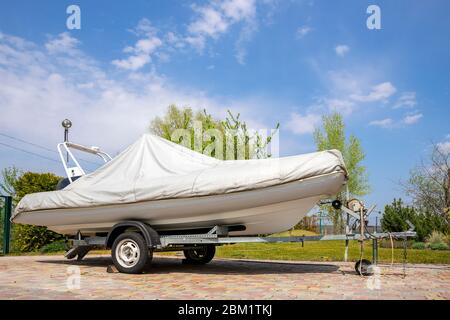 Großes modernes aufblasbares Motorboot-Schiff mit grauen oder weißen Schutztarpe auf Stahl-Sattelauflieger zu Hause Hinterhof an hellen sonnigen Tag bedeckt Stockfoto
