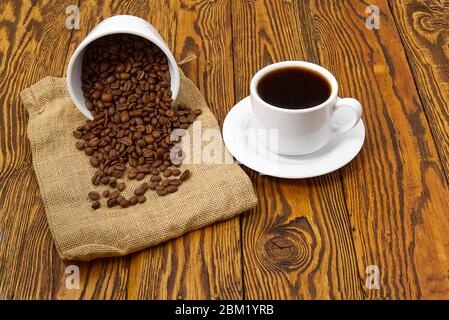 Weiße Kaffeetasse mit aromatischem Kaffeegetränk auf braunem Holztisch. Kaffeesamen auf antikem Holztisch mit Tasse Kaffee Getränk Stockfoto