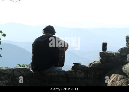 Junger Mann, der in den Bergen Gitarre spielt, mit natürlichem Panorama vor ihm Stockfoto