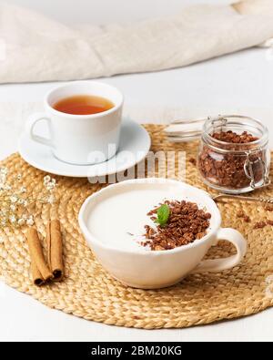 Joghurt mit Schokoladengranola in Tasse, Frühstück mit Tee auf beigem Hintergrund, Seitenansicht, vertikal. Stockfoto