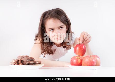 Kinder und Schokolade. Ein fröhliches Mädchen wählt zwischen Schokolade und Äpfeln. Gesunde Ernährung Stockfoto