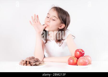 Kinder und Schokolade. Ein fröhliches Mädchen wählt zwischen Schokolade und Äpfeln. Gesunde Ernährung Stockfoto