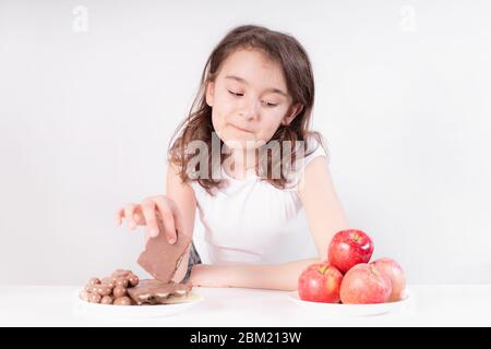 Kinder und Schokolade. Ein fröhliches Mädchen wählt zwischen Schokolade und Äpfeln. Gesunde Ernährung Stockfoto