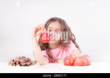 Kinder und Schokolade. Ein fröhliches Mädchen wählt zwischen Schokolade und Äpfeln. Gesunde Ernährung Stockfoto