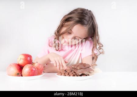 Kinder und Schokolade. Ein fröhliches Mädchen wählt zwischen Schokolade und Äpfeln. Gesunde Ernährung Stockfoto