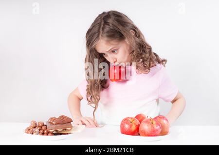 Kinder und Schokolade. Ein fröhliches Mädchen wählt zwischen Schokolade und Äpfeln. Gesunde Ernährung Stockfoto