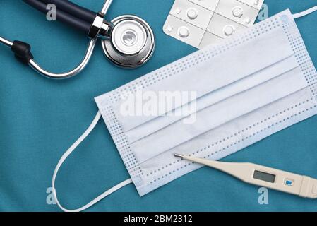 Hochwinkelfoto einer chirurgischen / Covid-19 Maske, einem Stethoskop, Digitalthermometer und Pillenpack auf grünem Tuch chirurgische Peelings. Stockfoto