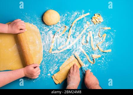 Paar bereitet italienische Pasta von Hand auf blauem Hintergrund. Flach legen mit Pasta machen. Rollender Grieß gelber Teig. Formen Tagliatelle Pasta. Stockfoto