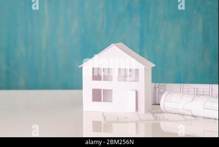Weißes Modellhaus mit Bauplänen auf einer reflektierenden Oberfläche vor türkisfarbenem Hintergrund Stockfoto
