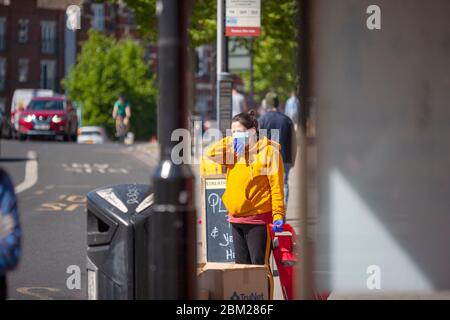 Frau stand an einer Londoner Bushaltestelle mit persönlicher Schutzausrüstung und berührte ihr Gesicht. Streatham High Road, Lambeth, London, Großbritannien Stockfoto
