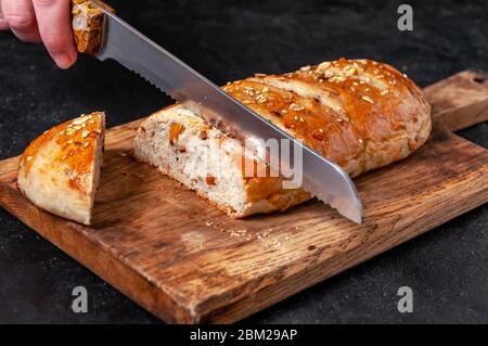 Frau schneidet selbstgemachte Vollkornbrot mit Leinsamen und Sesam auf Holzbrett auf Dark Table Stockfoto