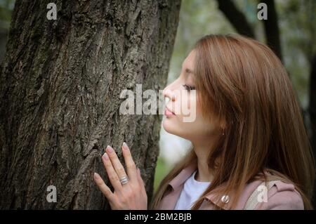 Eine junge attraktive Frau mit blondem Haar posiert in der Nähe eines großen Baumes in einem Frühlingspark im Freien. Sie lehnt sich an einen Baum, um die Rinde zu berühren Stockfoto