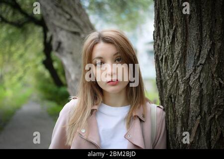 Outdoor weibliche Porträt der jungen attraktiven Frau mit einem blonden Haar posiert in der Nähe eines großen Baumes in einem Frühlingspark im Freien. Sie lehnt sich an einen Baum an Stockfoto