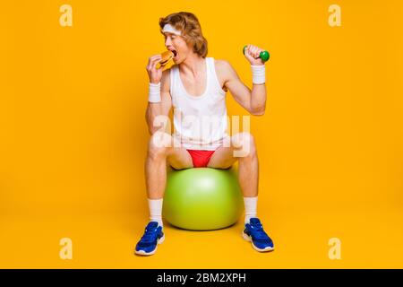 Porträt von seinem er schön attraktiv funky schlank dünnen Kerl sitzt auf fitball Essen lecker lecker lecker schnelles Essen tun Arbeit aus leicht Hantel isoliert über Stockfoto