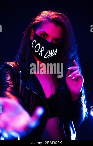 Porträt eines winkenden Attraktive Mädchen trägt schwarze Schutzmaske mit Corona Zeichen auf Gesicht in Neon Lights mit schwarzem Foggy Hintergrund Stockfoto