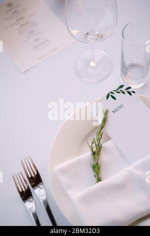 Empfang am Tisch für Hochzeitsessen. Weißer Teller mit Tuch Handtuch mit Papier-Menü und Zweig Rosmarin, vor dem Hintergrund der weißen Tischdecke Stockfoto