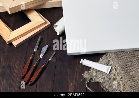 Eine weiße leere Leinwand auf Trage, Palettenmesser, Unterrahmen, ein Rohr mit Öl- oder Acrylfarbe und Sacktuch, das auf einem braunen Holztisch liegt Stockfoto