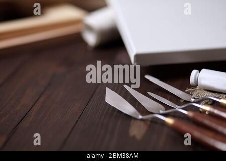 Palettenmesser liegen auf einem braunen Holztisch. Selektiver Fokus. Zweiter Plan: Weiße leere Leinwand und Tragbahren auf unscharfem Hintergrund Stockfoto