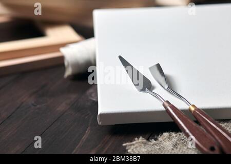 Künstler-Werkzeuge: Palettenmesser liegen auf einer weiß grundierten Baumwollleinwand. Holztragen im verschwommenen Hintergrund. Selektiver Fokus Stockfoto